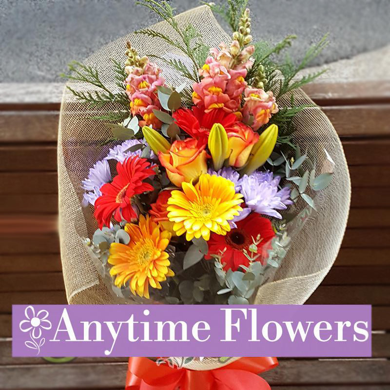 A vibrant bouquet with orange roses, golden gerberas, red gerberas, purple asters, and snapdragons.