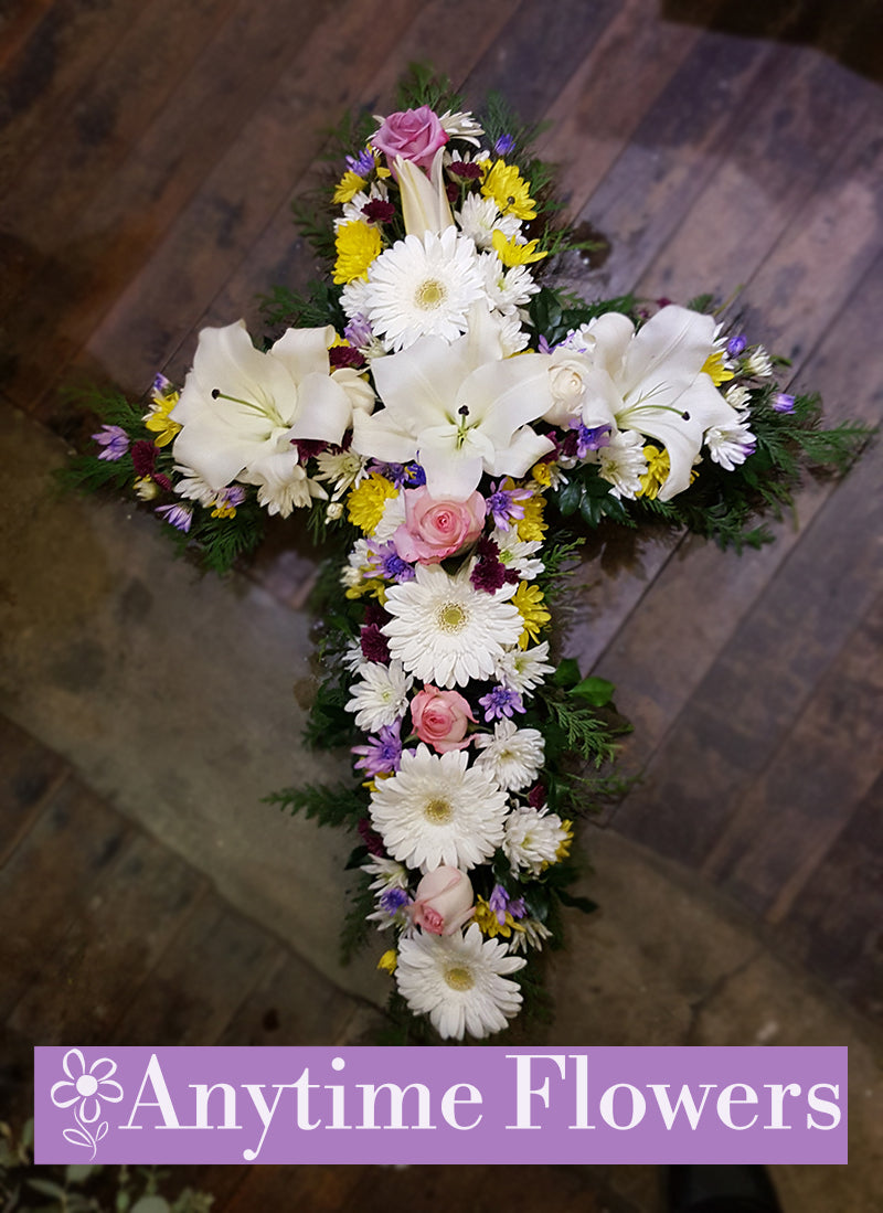 A floral cross arrangement featuring white lilies, gerberas, pink roses, and purple asters.