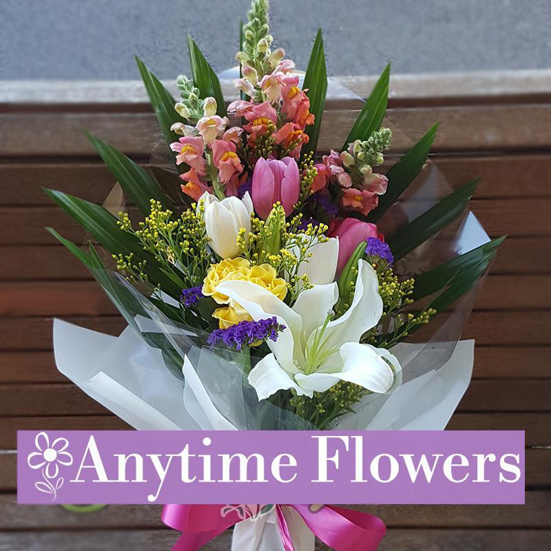 A cheerful bouquet of pink tulips, white lilies, yellow roses, and purple statice, accented with greenery and tied with a pink ribbon.