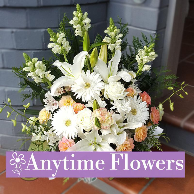 A graceful arrangement of white gerberas, lilies, cream roses, and peach carnations, accented with greenery and displayed in a white vase.