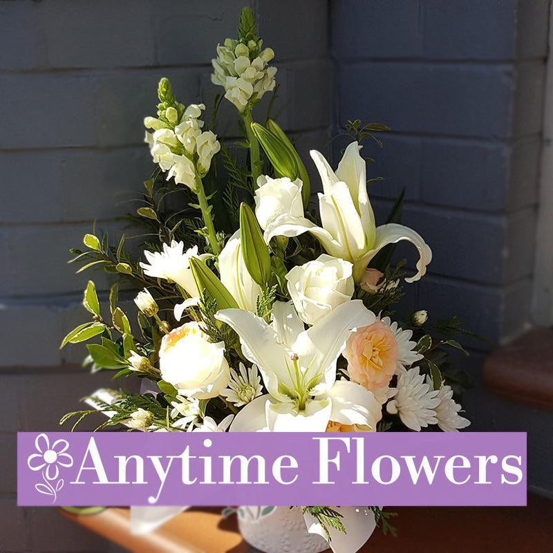 A classic white centerpiece featuring lilies, roses, and baby's breath, accented with greenery and arranged in a stylish vase.