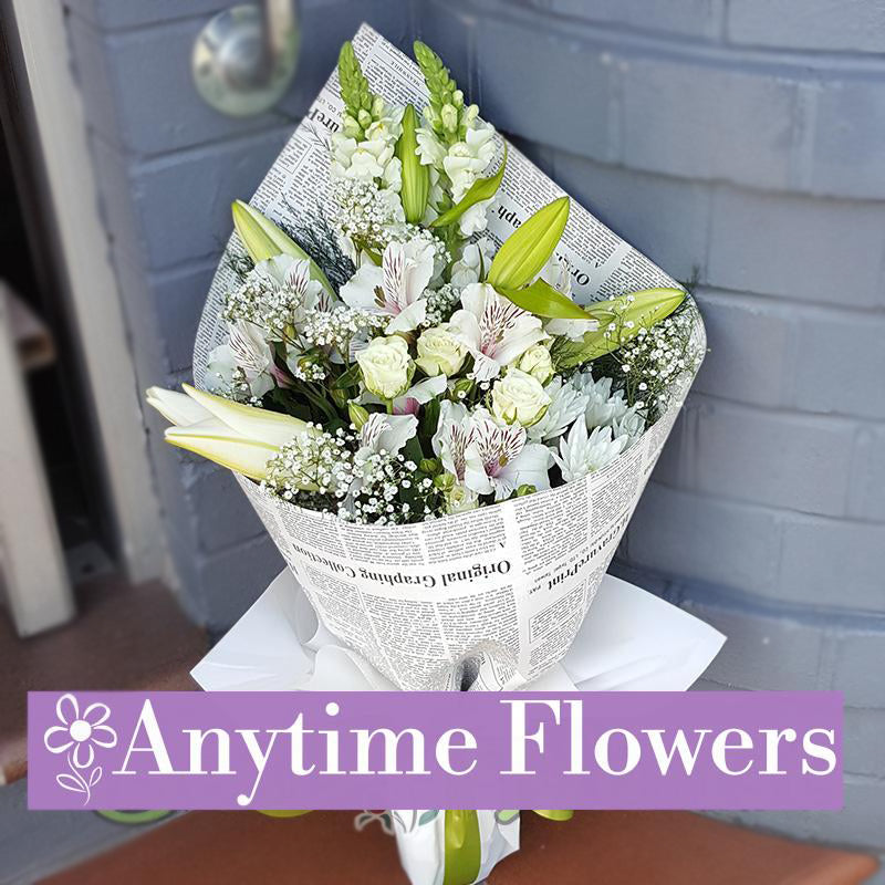 A classic white bouquet featuring lilies, roses, and baby's breath, accented with lush greenery and wrapped in elegant newsprint.