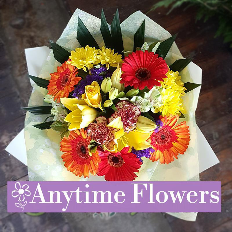A vibrant bouquet featuring yellow lilies, red and orange gerberas, purple statice, and carnations, accented with lush greenery for a joyful design.
