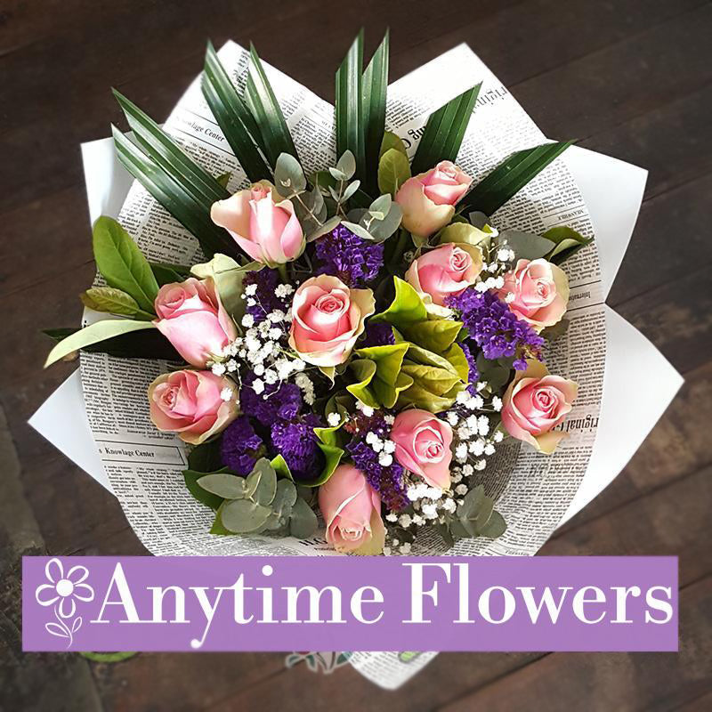 A stylish bouquet featuring pink roses, purple statice, and baby's breath, wrapped in newsprint and accented with lush greenery for a sophisticated touch.