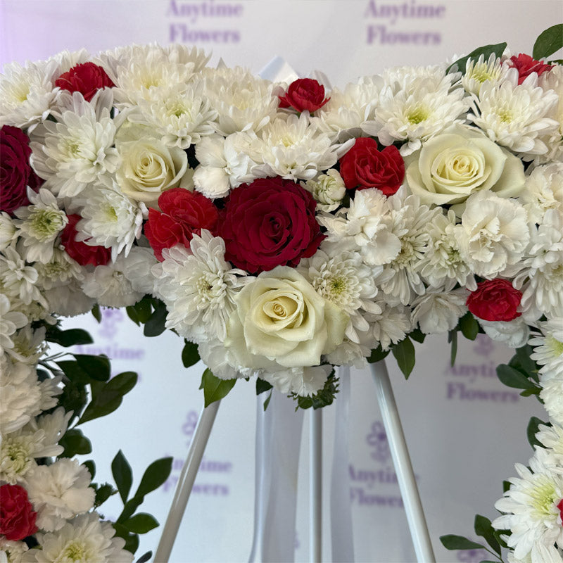 A heart-shaped floral wreath featuring white chrysanthemums, red roses, and white roses, elegantly arranged with lush greenery for a meaningful tribute.