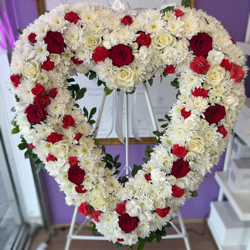 A close-up of a premium heart-shaped sympathy wreath with delicate white chrysanthemums, deep red roses, and soft white roses.
