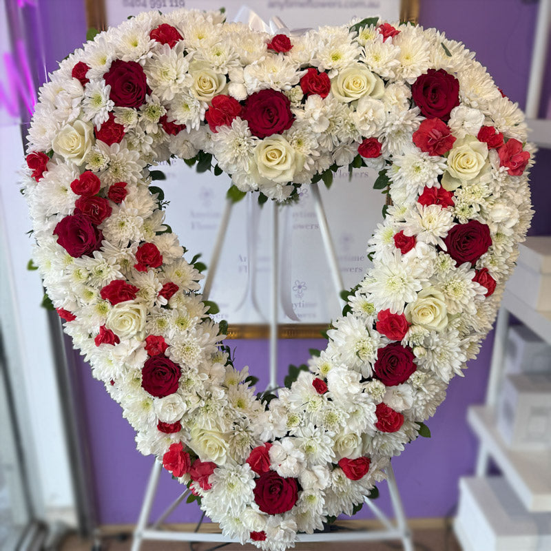 A full view of a heart-shaped memorial wreath, featuring fresh white and red flowers, presented on a stand with a satin ribbon for a graceful tribute.