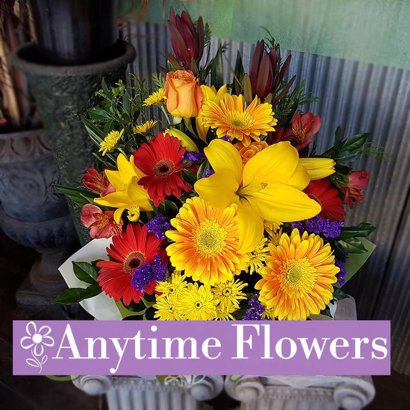 A vibrant arrangement of yellow and red gerberas, orange roses, and purple statice with lush greenery.
