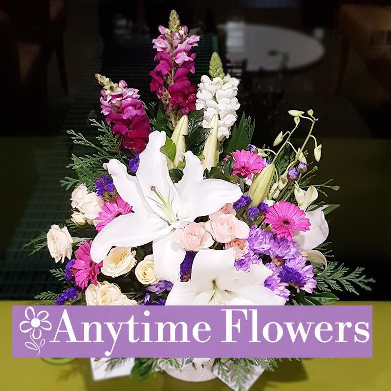 Elegant centerpiece featuring white lilies, pink roses, and purple flowers with lush greenery.