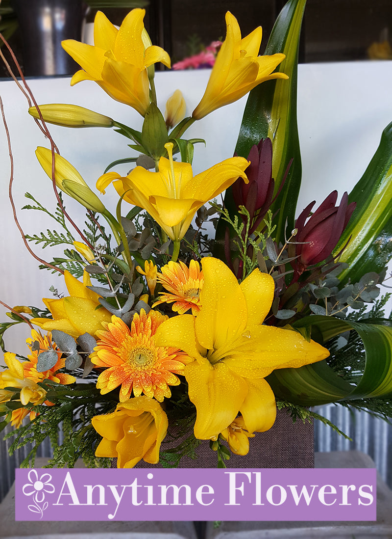 Bright arrangement of yellow lilies and gerberas with greenery in a box.