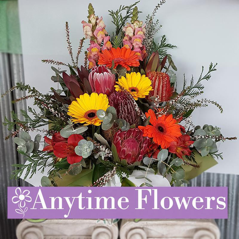 A vibrant bouquet of orange and yellow gerberas, proteas, and snapdragons with lush greenery.