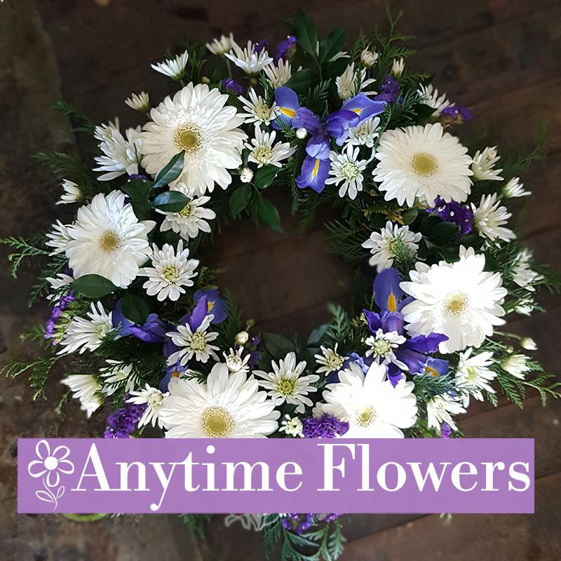 A delicate wreath with white gerberas, daisies, and purple irises, accented by lush greenery.