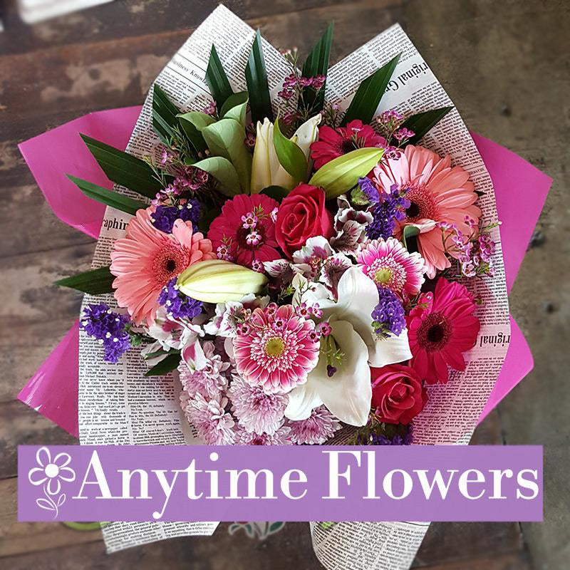 A vibrant bouquet featuring pink gerberas, roses, lilies, and purple statice wrapped in stylish paper.