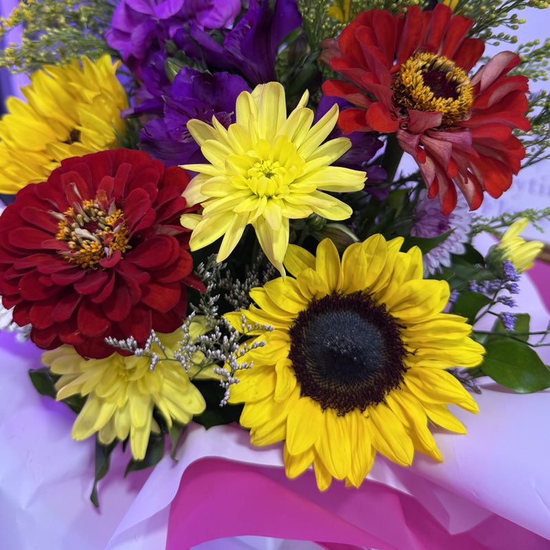 A cheerful mini-box arrangement of yellow sunflowers, purple alstroemeria, and other bright blooms, wrapped in soft white tissue paper.