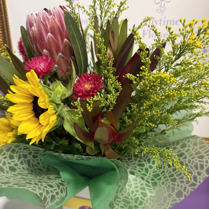 A native flower mini-box with a striking combination of proteas, sunflowers, and vibrant red blossoms, wrapped in tissue paper.