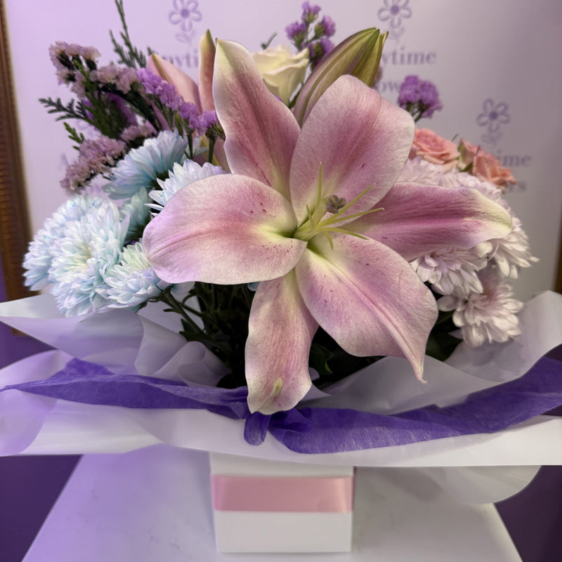 A delicate mini-box arrangement featuring soft pink lilies, lavender chrysanthemums, and roses, wrapped in purple tissue paper.