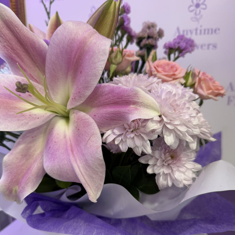A soft and beautiful mini-box arrangement of pink lilies, pink roses, and purple flowers, perfect for a thoughtful gift.