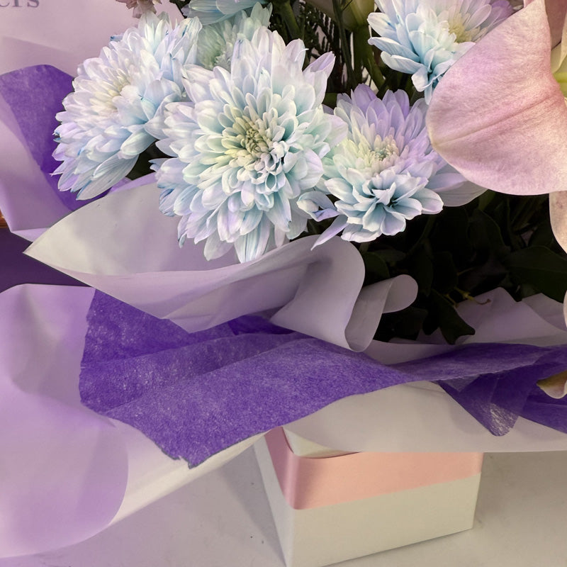 A stunning floral mini-box featuring a combination of soft pink lilies, pink roses, and purple flowers, wrapped in delicate tissue paper.