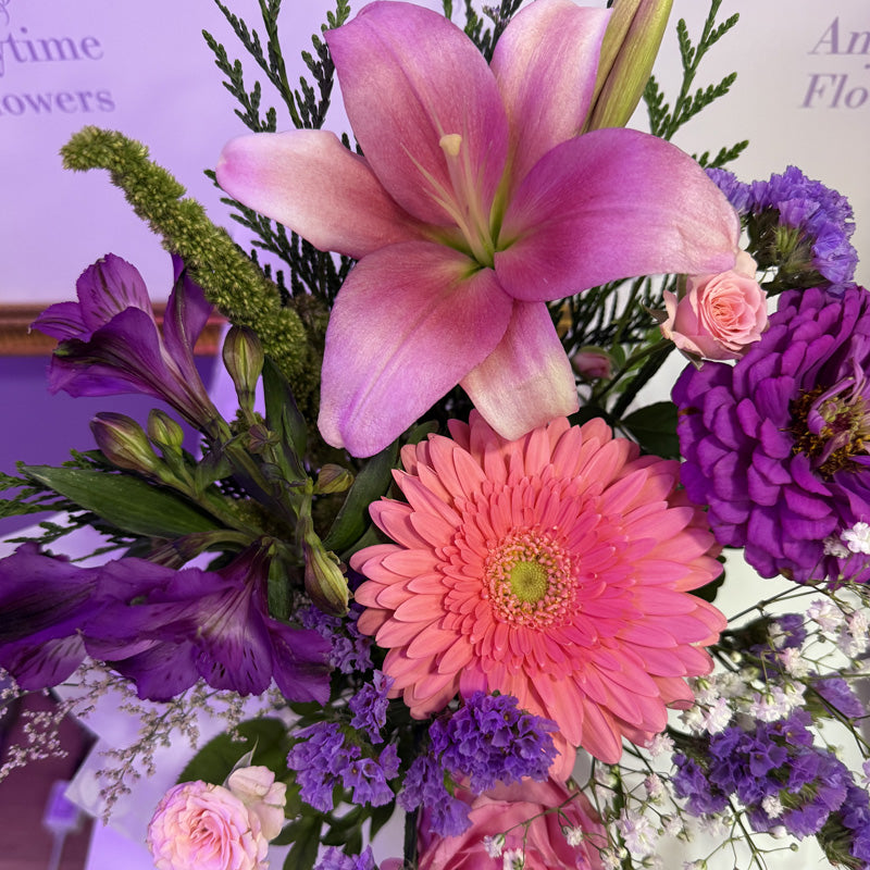 A mini-box arrangement featuring soft pink lilies, purple chrysanthemums, and pink roses, wrapped in purple tissue paper for a delicate, elegant touch.