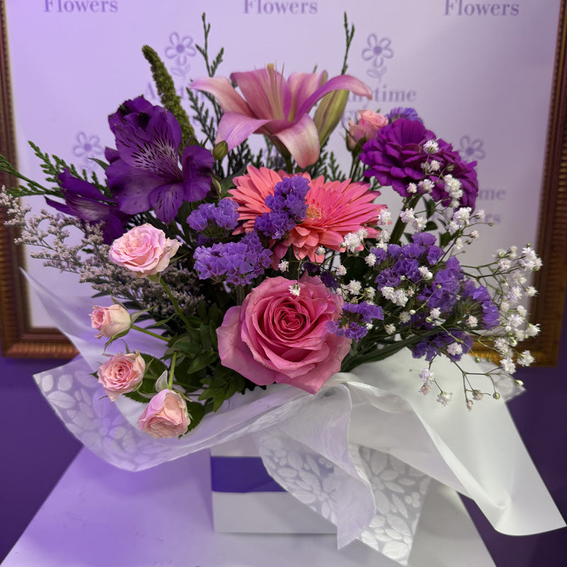 A soft and beautiful mini-box arrangement of pink lilies, pink roses, and purple flowers, perfect for a thoughtful gift.