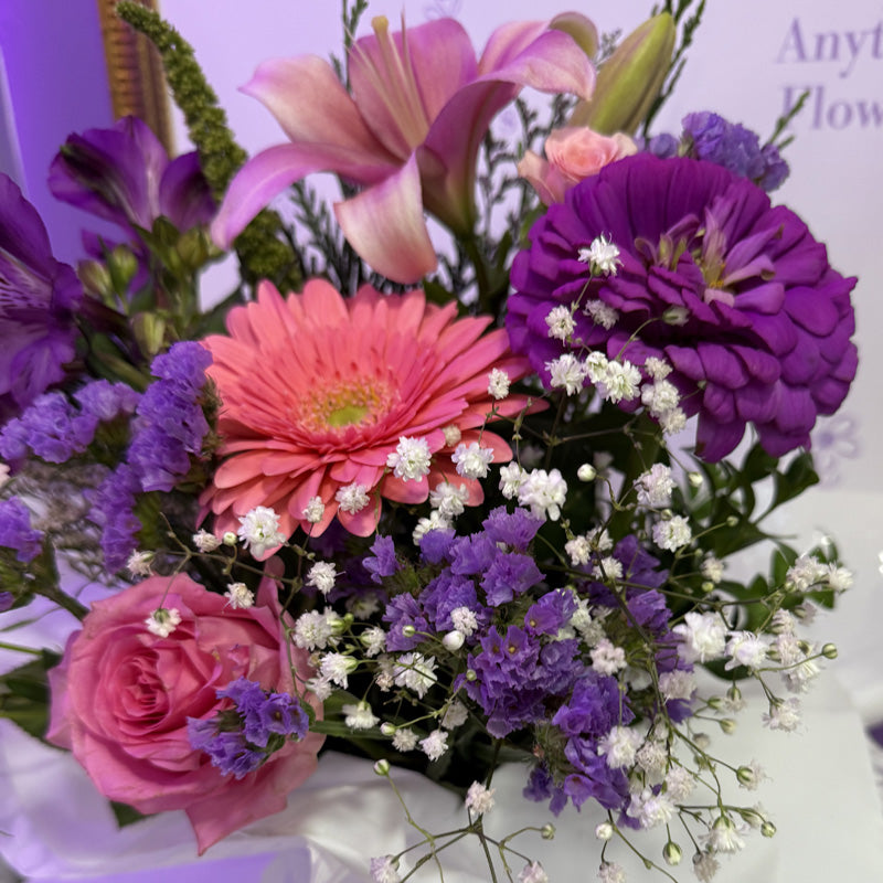 A stunning floral mini-box featuring a combination of soft pink lilies, pink roses, and purple flowers, wrapped in delicate tissue paper.