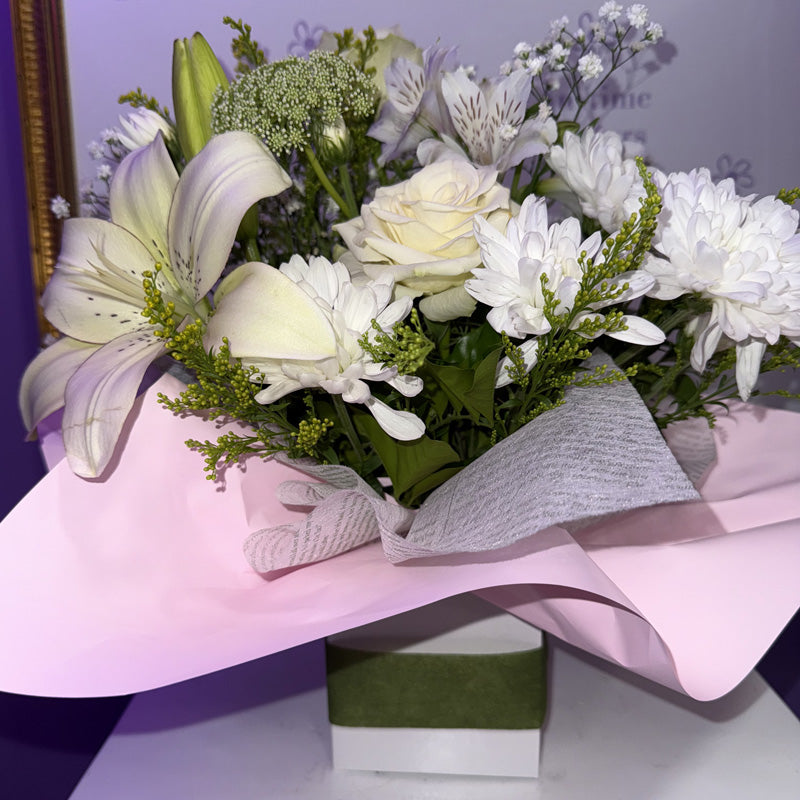 Close-up of white lilies, roses, and chrysanthemums with fresh green accents, placed in a mini box.