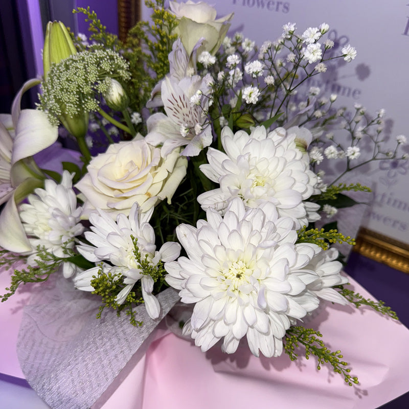 A white flower arrangement featuring lilies, roses, and delicate greenery wrapped in soft gray tissue paper.