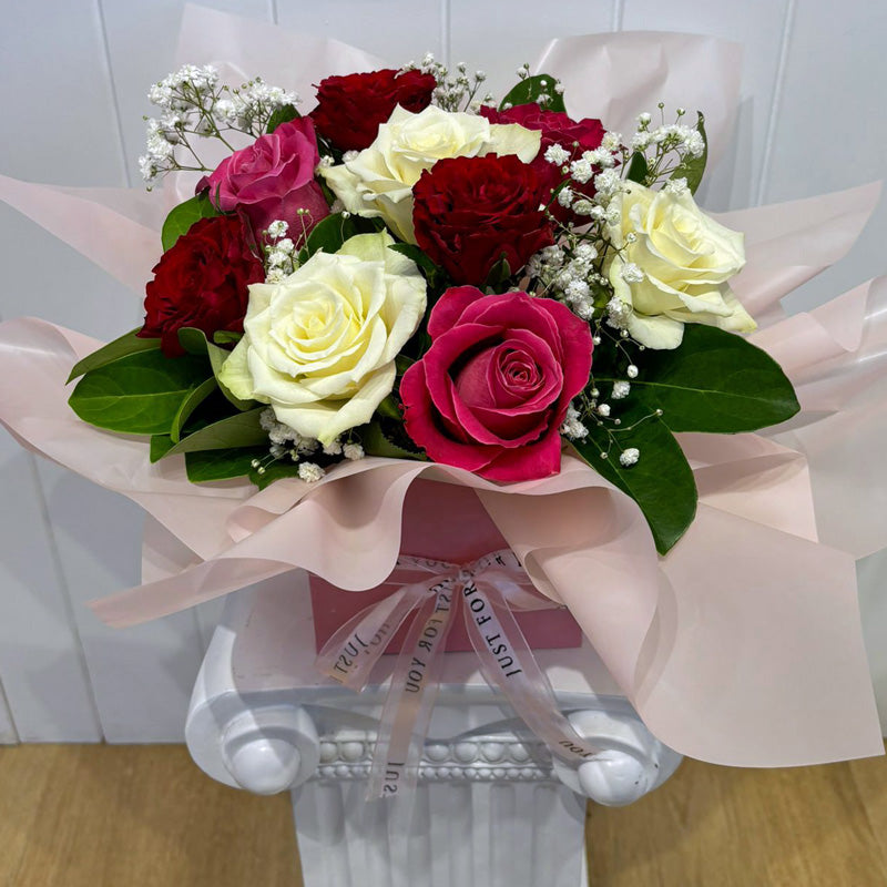 A beautifully arranged floral box featuring red, white, and pink roses, accented with baby’s breath and lush greenery, wrapped in soft pink tissue.